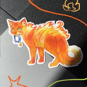 Op de afbeelding: Een gestanste sticker van een oranje kat met vacht die overgaat in gestileerde vlammen. De kat heeft groene ogen en houdt een kleine blauwe halsband met een gouden belletje in de bek. De illustratie bevat levendige tinten geel, oranje en rood.