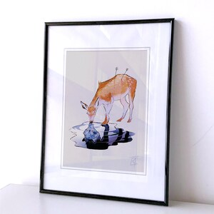 May include: Framed art print featuring a watercolor illustration of a deer drinking from a pool of water. The deer is depicted in shades of brown and orange, with arrows in its back. The water reflects a distorted image of a figure.
