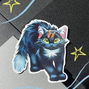 Op de afbeelding: Een gestanste sticker met een donkerblauwe, pluizige kat met grote, heldergroene ogen en een kleine oranje vlinder op het voorhoofd. De kat heeft een witte vlek op de borst en een lichtblauwe staartpunt, tegen een donkere achtergrond met sterren.