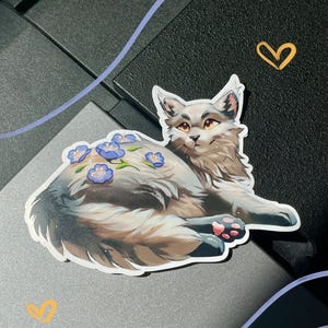 Op de afbeelding: Een gestanste sticker van een langharige kat met lichtbruine en cr&egrave;mekleurige vacht, amberkleurige ogen en roze voetzooltjes. De kat ligt neer met verschillende kleine blauwe bloemen met gele hartjes op de rug, in een digitale kunststijl.