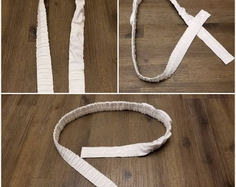 May include: White elastic band with a snap closure. The band is shown in three different positions, demonstrating how it can be used to hold a garment in place.