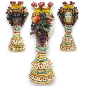 Puede incluir: Tres jarrones de cerámica con forma de cabeza, coronas ornamentadas, frutas esculpidas y racimos de uvas. Dos jarrones muestran rostros con tonos de piel distintos, mientras que el central muestra un perfil lateral. Base con rayas pintadas.