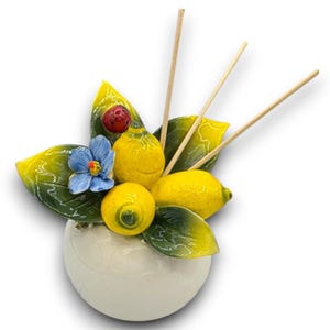 Pode incluir: Uma base de difusor de cerâmica branca com um arranjo decorativo de dois limões amarelos, uma flor azul, uma joaninha vermelha e folhas verdes. Três varetas de madeira finas estendem-se para cima a partir do centro do conjunto cerâmico.