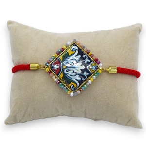 Puede incluir: Pulsera de cuerda roja con un dije cuadrado que presenta un diseño de azulejos coloridos e intrincados. El dije está rodeado de pequeñas cuentas de colores.