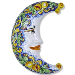 Peut inclure: Une plaque murale en forme de croissant de lune en céramique avec une finition craquelée brillante. Le visage est blanc avec des lèvres brunes et des yeux sombres. Le bord extérieur présente un motif peint à la main de grenades jaunes.