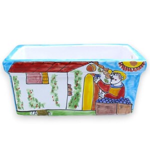 Op de afbeelding: Een rechthoekige keramische plantenbak met een handgeschilderde scène van een wit huis met een rood dak, groene klimplanten en een persoon met een gele hoed die blauwe druiven oogst in houten kratten. De rand is fel lichtblauw gekleurd.