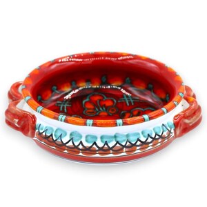Puede incluir: Un cuenco redondo de cerámica con dos asas laterales, que presenta un interior y un exterior de color rojo vibrante. El diseño incluye patrones pintados a mano en turquesa y negro, con una banda blanca en el medio y un borde decorativo naranja y rojo.