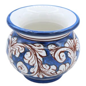 May include: A small, round ceramic pot featuring a white interior and a blue rim. The exterior is decorated with a hand-painted pattern of white and brown swirling acanthus leaf motifs set against a vibrant blue background, creating a distinct contrast.