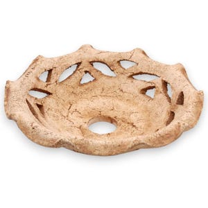 May include: A shallow, circular ceramic bowl with a textured, light brown finish. The rim features a scalloped edge with a series of triangular cutouts arranged in a pattern around the perimeter, and a single circular hole is centered at the base.