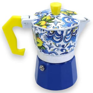 Puede incluir: Una cafetera italiana con base azul sólida y cuerpo superior blanco decorado con un patrón floral azul y limones amarillos. El dispositivo incluye un asa de color amarillo brillante y un pomo a juego en la tapa.