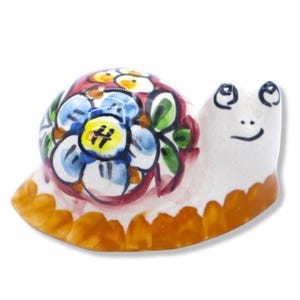 Può includere: Una statuetta di lumaca in ceramica con corpo bianco e finitura lucida. Il guscio presenta un motivo floreale dipinto a mano nei toni del blu, giallo e verde, delineato in rosso scuro. La base della lumaca è decorata con un bordo arancione smerlato.