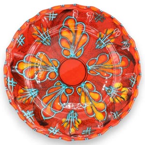 Puede incluir: Un plato de cerámica redondo con borde festoneado y un color base rojo vibrante. La superficie está decorada con un círculo rojo central rodeado por seis motivos estilizados en forma de hoja en naranja y amarillo, delineados en azul turquesa.