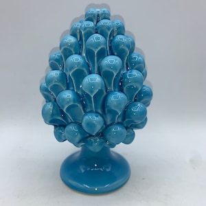 Puede incluir: Un adorno de piña de cerámica azul con un acabado liso y brillante. La piña está detallada con escamas individuales y se asienta sobre una base redonda.