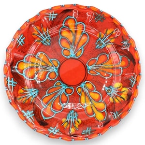 Puede incluir: Un plato de cerámica redondo con borde festoneado y un color base rojo vibrante. La superficie está decorada con un círculo rojo central rodeado por seis motivos estilizados en forma de hoja en naranja y amarillo, delineados en azul turquesa.