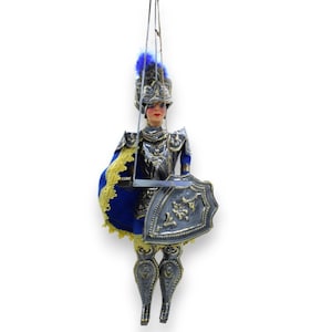 Può includere: Un burattino di un cavaliere che indossa un'armatura argentata con un mantello blu e oro, che tiene uno scudo argentato con accenti dorati. Il burattino ha un viso dipinto con labbra rosse e occhi blu.