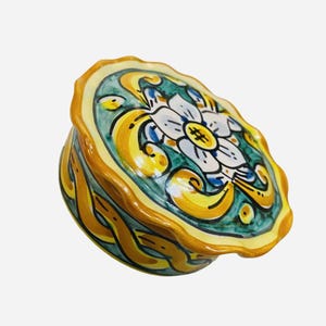 Puede incluir: Un joyero redondo de cerámica con tapa de borde festoneado. La tapa presenta una flor blanca pintada a mano con centros amarillos sobre un fondo verde azulado, rodeada de volutas amarillas. La base tiene un patrón trenzado amarillo y verde azulado.