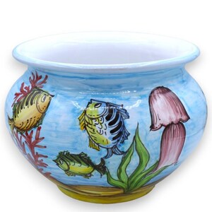 Op de afbeelding: Een ronde keramische plantenpot met een witte binnenkant en een lichtblauwe buitenkant met handgeschilderde onderwaterscènes. Het ontwerp bevat geel-blauw gestreepte vissen, een groene vis, rood koraal, groen zeewier en een roze kwal.