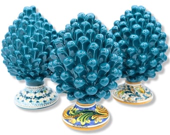 Puede incluir: Tres esculturas de piñas de cerámica en color verde azulado con acabados brillantes. Cada pieza presenta un diseño texturizado de escamas superpuestas y se asienta sobre una base circular decorada con motivos florales pintados a mano.