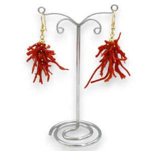 May include: A pair of dangling earrings featuring clusters of bright red, branch-like coral pieces suspended from gold-toned hooks. The earrings are displayed on a silver-toned metal stand with a spiral base and two curved arms for hanging jewelry.
