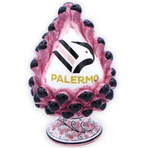 May include: A pink ceramic pine cone ornament with black details and a white eagle logo with the word "PALERMO" in gold lettering. The base of the ornament is decorated with a pink and white floral pattern.