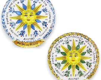 Può includere: Due piatti rotondi in ceramica con sfondo bianco e un sole giallo con un viso al centro. Il sole ha otto raggi ed è circondato da un cerchio di testo in italiano. Il testo è in nero e recita "ROSA dei VENTI".