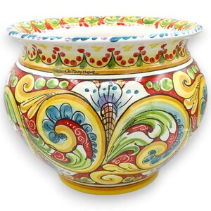 May include: A ceramic planter featuring a wide, flared rim and a rounded body. The exterior is hand-painted with vibrant yellow, red, green, and blue floral scrollwork patterns. The text Caltagirone is visible on the upper section of the pot.