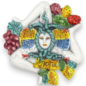 Puede incluir: Una Trinacria de cerámica blanca, símbolo de Sicilia, con una cabeza de Medusa con serpientes, alas coloridas y frutas. El diseño incluye uvas, higos chumbos y limones. La palabra "Sicilia" está escrita en una pancarta.