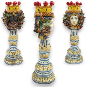 Puede incluir: Tres candelabros de cerámica con cabezas esculpidas que llevan coronas amarillas con detalles rojos. Cada base está pintada con rayas azules y blancas. Las cabezas están adornadas con frutas coloridas y racimos de uvas, mostrando rasgos faciales detallados.