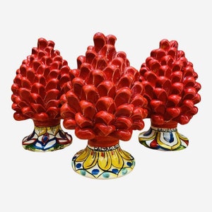 Op de afbeelding: Drie rode keramische dennenappelsculpturen met kleurrijk beschilderde bases. De bases dragen de woorden "Caltagirone" en "Sicilië".