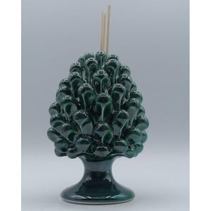 Op de afbeelding: Een donkergroene keramische diffuser in de vorm van een dennenappel met een glanzende afwerking. De diffuser heeft een voetstuk en bevat verschillende houten stokjes. Het dennenappelontwerp is gedetailleerd en gestructureerd.