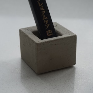 Può includere: Un piccolo portapenne in cemento grigio con una matita nera all'interno. La matita ha la scritta giapponese "Premium Quality 62".