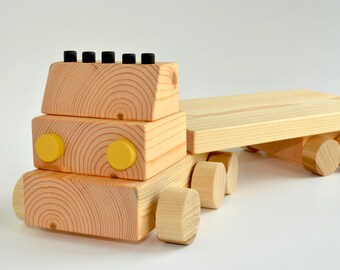 May include: A wooden toy truck with a flatbed trailer, featuring a natural light wood grain finish, two round yellow headlights, and five small black cylindrical accents on the roof of the cab. The vehicle has multiple round wooden wheels.