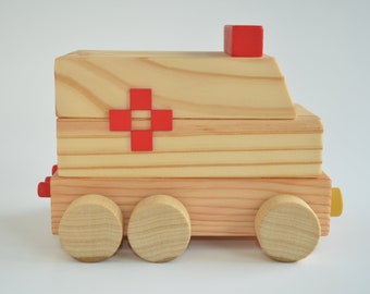 May include: A wooden toy ambulance featuring a natural light wood grain finish, three circular wheels on the side, a red cross symbol on the center panel, and a small red block on the roof. The vehicle has a yellow accent piece at the rear.