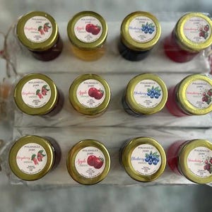 May include: An overhead shot of twelve small jars of jam, each with a gold lid and a label featuring fruit illustrations. The flavors include raspberry, apple, blueberry, and strawberry. The jars are arranged in rows on white rectangular trays.