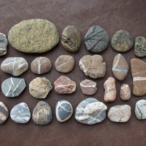 May include: A collection of 25 smooth, flat, gray, white, and brown stones with various patterns. The stones are arranged in rows on a brown surface.