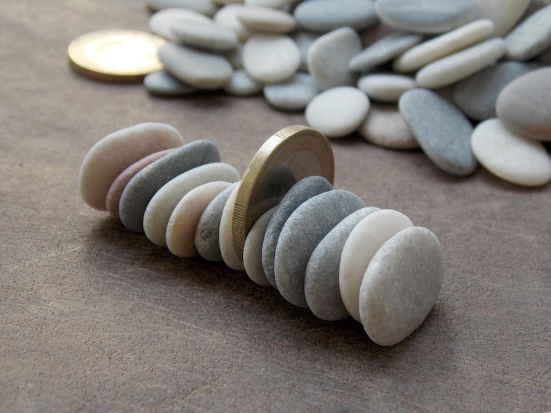 May include: A horizontal row of smooth, rounded river stones in shades of white, gray, and tan, arranged upright and interspersed with a single gold-colored coin standing vertically among them, with a pile of loose stones in the background.