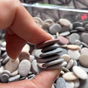 May include: A stack of six small, smooth, oval-shaped river stones held between fingers. The stones vary in color, including shades of gray, light brown, and muted pink. Below the stack is a large collection of similar loose river stones.