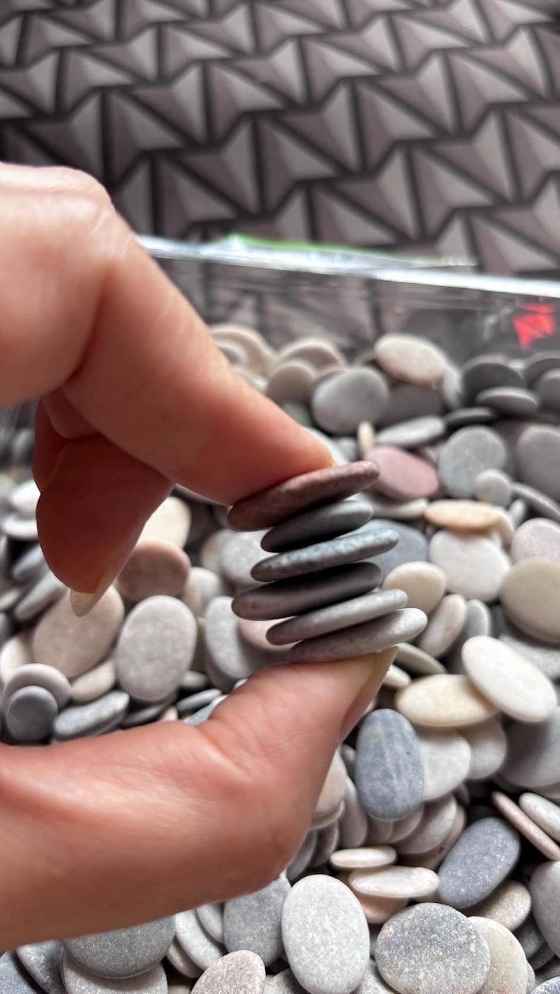 May include: A stack of six small, smooth, oval-shaped river stones held between fingers. The stones vary in color, including shades of gray, light brown, and muted pink. Below the stack is a large collection of similar loose river stones.