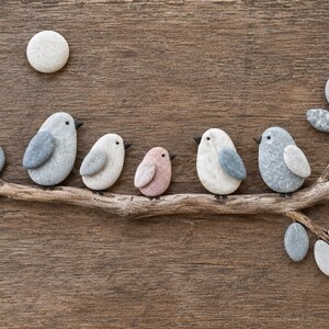 May include: A wall art piece featuring five birds made of smooth, rounded stones perched on a driftwood branch. The birds are gray, white, and light pink with small black beaks. A single white stone moon sits above the branch.