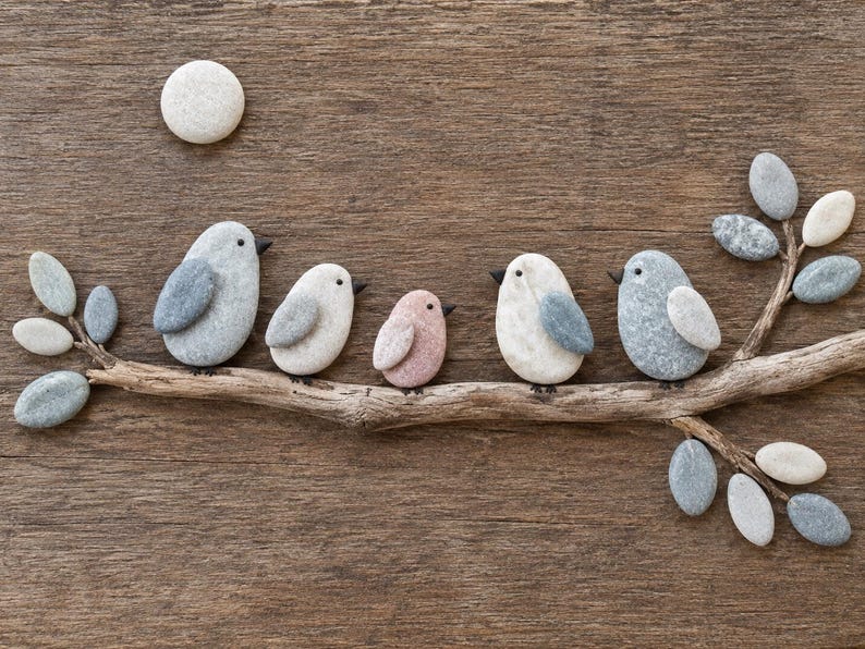 May include: A wall art piece featuring five birds made of smooth, rounded stones perched on a driftwood branch. The birds are gray, white, and light pink with small black beaks. A single white stone moon sits above the branch.