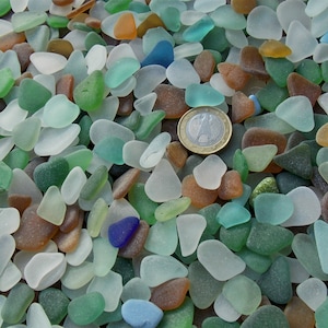 Puede incluir: Una colección de piezas de vidrio marino lisas y desgastadas en varios tonos de blanco, verde, marrón y azul, esparcidas. Una moneda de un euro está colocada entre los fragmentos de vidrio para proporcionar una referencia de escala de las piezas.