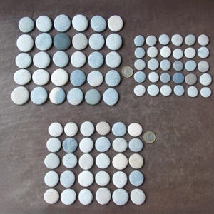 May include: Arrangement of smooth, flat, round stones in varying shades of blue, gray, and white. The stones are organized in rectangular patterns on a brown surface. A coin is placed near some of the stone arrangements.
