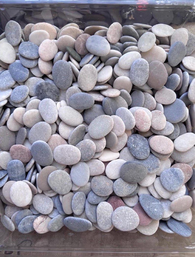 May include: A collection of numerous small, smooth, oval-shaped river stones in various shades of gray, white, beige, and light brown. The stones are packed closely together, displaying natural textures and rounded edges throughout the group.