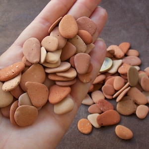 Puede incluir: Un montón de pequeñas piedras lisas de color terracota. Las piedras son de varios tonos de marrón y naranja, y algunas tienen un aspecto moteado.