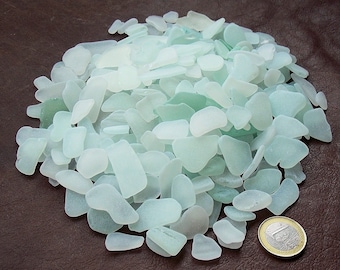 May include: A large collection of smooth, frosted sea glass pieces in various shades of pale aqua, mint green, and white. The pieces are irregular in shape and size, scattered in a pile next to a single circular gold and silver colored coin.