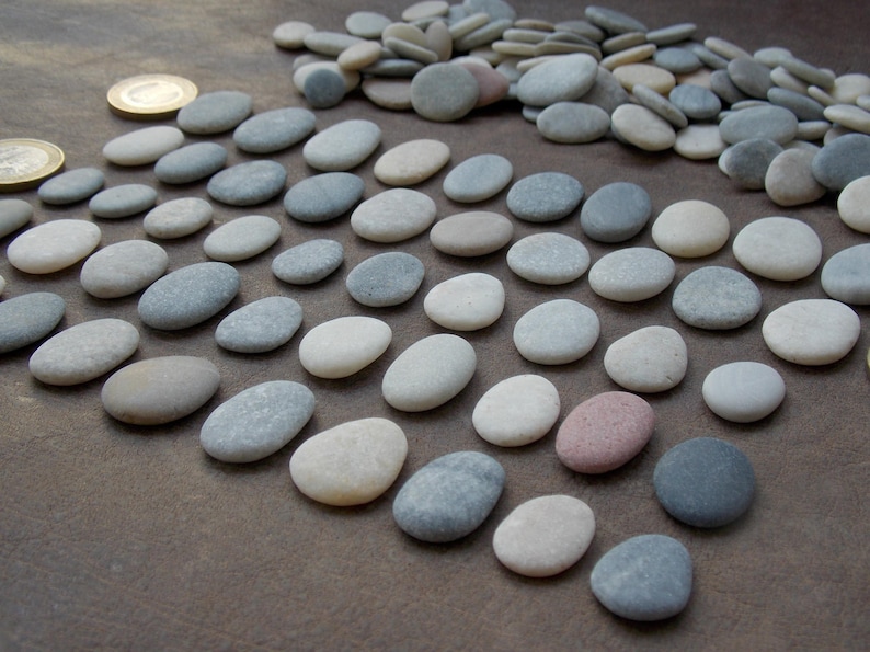 May include: A collection of smooth, rounded river stones in various shades of gray, white, and beige, arranged in a grid pattern with a pile of additional loose stones in the background and two gold-colored coins for scale.