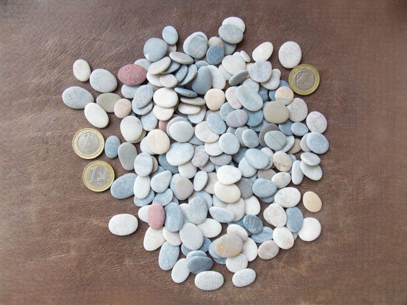 May include: A collection of small, smooth, oval-shaped river stones in shades of white, gray, blue, and tan, interspersed with three gold-colored euro coins for scale, scattered across a textured brown surface.