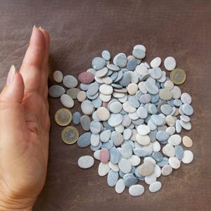 May include: A collection of small, smooth, oval-shaped river stones in shades of white, gray, blue, and tan, interspersed with three gold-colored coins for scale, arranged in a loose pile on a textured brown surface.