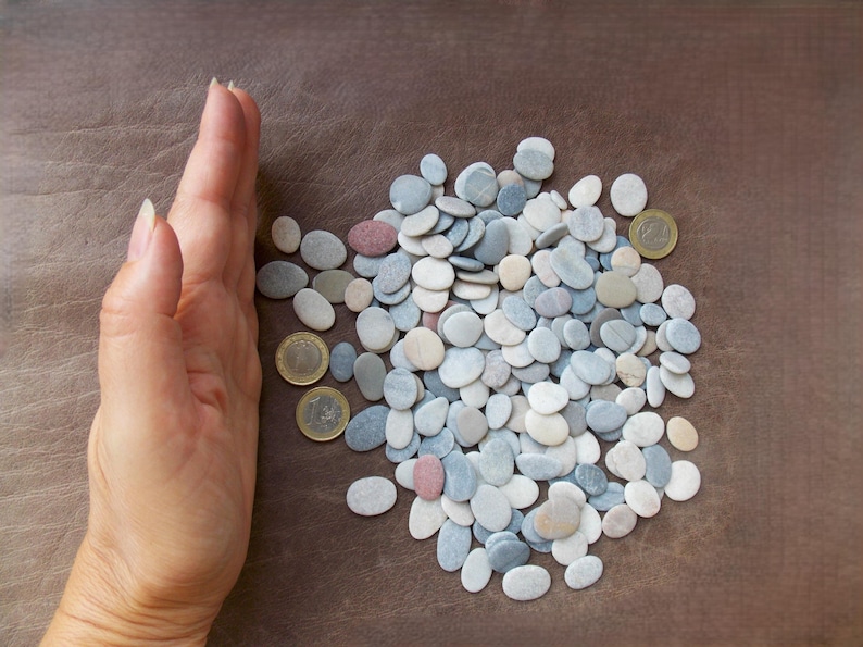 May include: A collection of small, smooth, oval-shaped river stones in shades of white, gray, blue, and tan, interspersed with three gold-colored coins for scale, arranged in a loose pile on a textured brown surface.