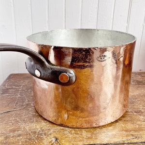 以下が含まれることがあります： 暗い色のハンドルとリベットが付いた銅製のソースパン。鍋の内側は光沢があり、反射性があり、外側はわずかにテクスチャ加工されています。鍋は木製の表面に置かれています。